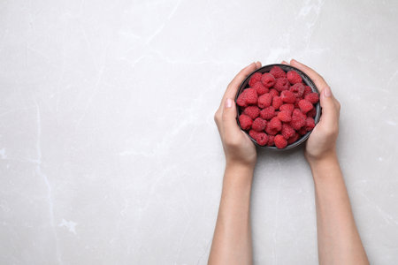 Woman holding bowl of delicious ripe raspberries on white table, top view. Space for text