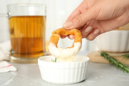 Woman dipping crunchy fried onion ring in sauce at gray marble table, closeupの写真素材