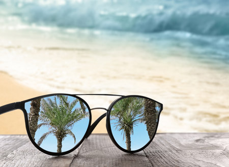 Palms mirroring in sunglasses on wooden desk at sandy beach near oceanの写真素材