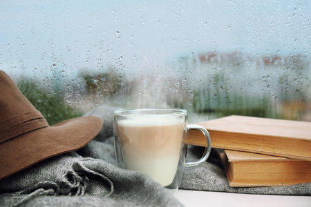 Cup of delicious hot drink, books and scarf on window sill after rainの写真素材