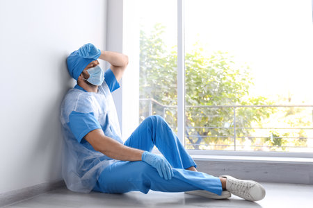 Exhausted doctor sitting on floor indoors. Stress of health care workers during COVID-19 pandemicの写真素材