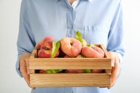 Woman holding wooden crate with fresh ripe donut peaches on light background, closeupの写真素材
