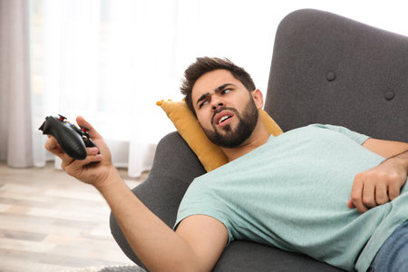 Lazy young man playing video game while lying on sofa at home
