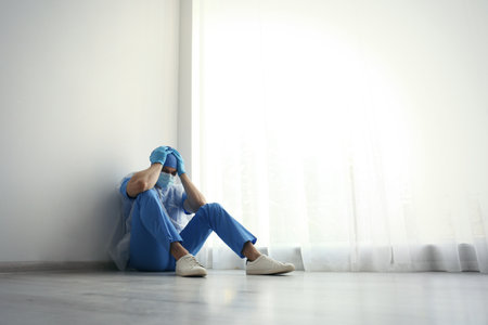 Exhausted doctor sitting on floor indoors, space for text. Stress of health care workers during COVID-19 pandemicの写真素材