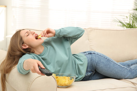 Lazy young woman with bowl of chips watching TV on sofa at homeの写真素材