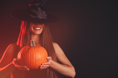 Young woman wearing witch costume with pumpkin on dark background, space for text. Halloween partyの写真素材