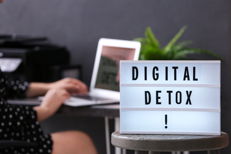 Woman using laptop at table in office, focus on lightbox with phrase DIGITAL DETOXの写真素材