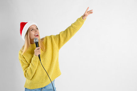 Emotional woman in Santa Claus hat singing with microphone on light gray background, space for text. christmas musicの写真素材