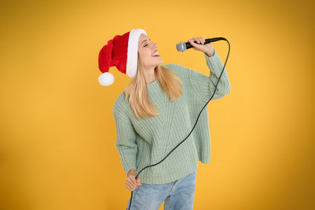Happy woman in Santa Claus hat singing with microphone on yellow background. christmas musicの写真素材