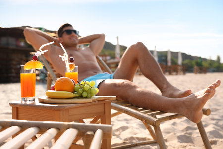 Happy man resting on sunny beach at resort, focus on table with drinks and fruitsの写真素材