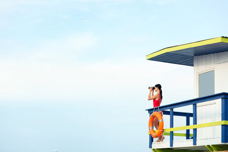 Female lifeguard with binocular on watch tower against blue skyの写真素材