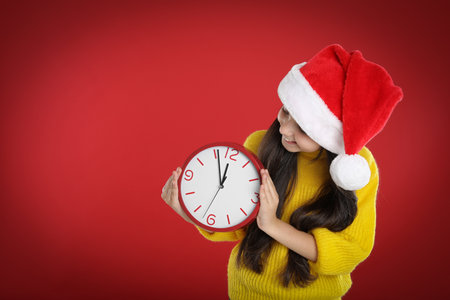 Girl in Santa hat with clock on red background, space for text. New Year count down
