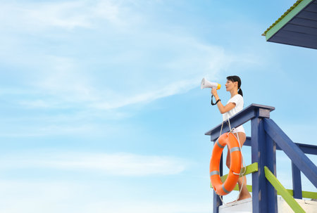 Female lifeguard with megaphone on watch tower against blue skyの写真素材