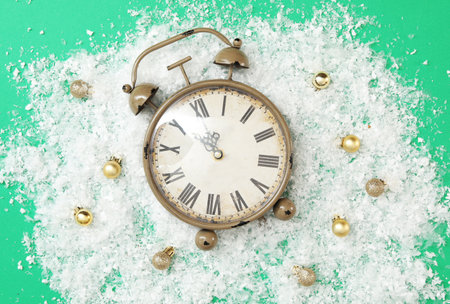 Alarm clock and festive decor in pile of snow on green background, flat lay. New Year countdown