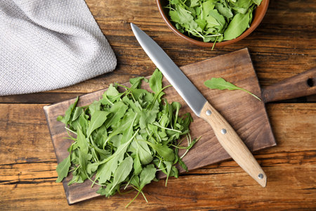 Cutting board with fresh arugula leaves and knife on wooden table, flat lay