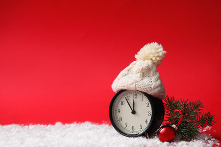 Alarm clock in hat and fir branch with Christmas ball on snow against red background, space for...