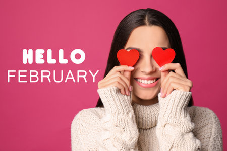 Greeting card with text Hello February. Young woman holding paper hearts near eyes on pink backgroundの写真素材