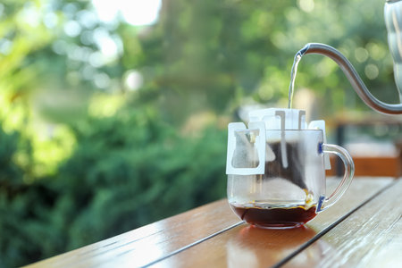 Pouring hot water into glass cup with drip coffee bag from kettle on wooden table outdoors. Space for textの写真素材
