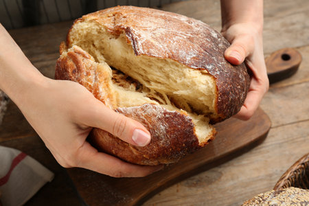 Woman breaking freshly baked bread at wooden table, closeup
