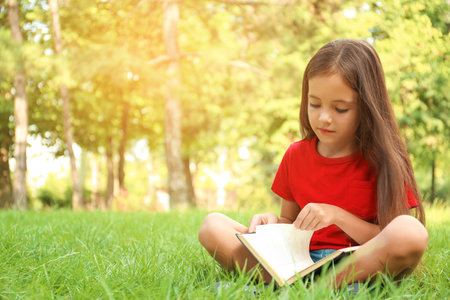Cute little girl reading book on green grass in parkの写真素材
