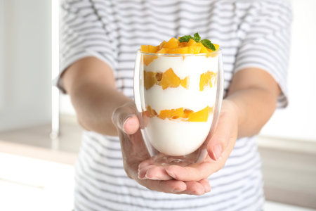 Woman holding glass of tasty peach dessert with yogurt in kitchen, closeupの写真素材