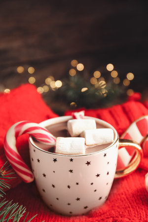 Cup of tasty cocoa with marshmallows and Christmas candy cane on knitted sweater against blurred festive lights. Space for textの写真素材