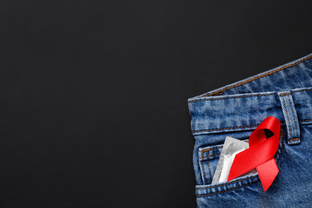 Top view of jeans with condom and red ribbon in pocket on black background, space for text. AIDS...