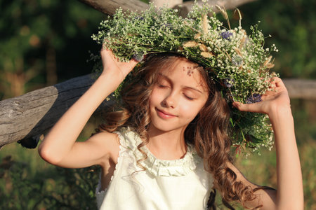 Cute little girl wearing wreath made of beautiful flowers near wooden fence outdoors on sunny dayの写真素材