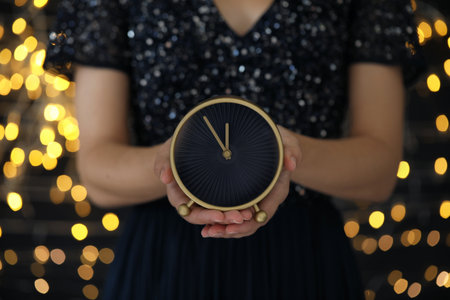 Woman holding alarm clock against blurred lights, closeup. New Year countdown