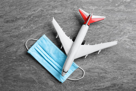 Toy airplane and medical mask on gray stone background, flat lay. Traveling during coronavirus...