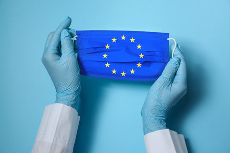 Doctor holding medical mask with European Union flag on light blue background, closeup. Coronavirus...