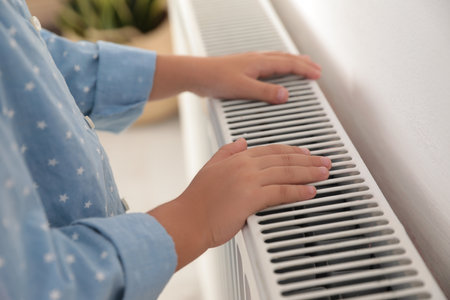 Child warming hands on heating radiator near white wall, closeup