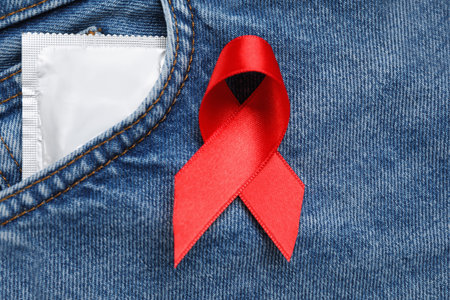 Jeans with red ribbon and condom, flat lay. AIDS disease awareness