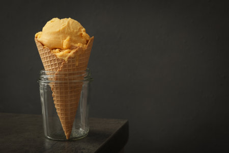 Delicious yellow ice cream in wafer cone and glass jar on gray table. Space for text