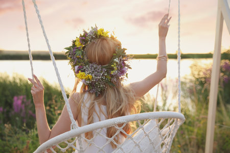 Young woman wearing wreath made of beautiful flowers on swing chair outdoors, back viewの写真素材
