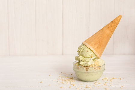 Delicious pistachio ice cream in wafer cone with chopped nuts on white wooden table. Space for textの写真素材