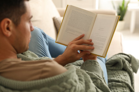 Man covered with warm green plaid reading book at home, closeupの写真素材