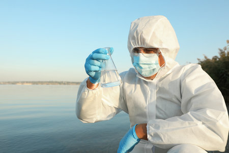 Scientist in chemical protective suit with conical flask taking sample from river for analysisの写真素材