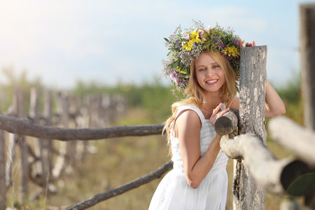 Young woman wearing wreath made of beautiful flowers near wooden fence on sunny dayの写真素材