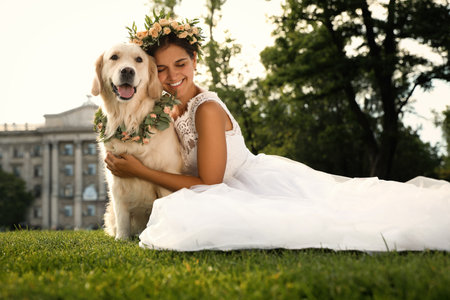 Bride and adorable Golden Retriever wearing wreath made of beautiful flowers on green grass outdoorsの写真素材
