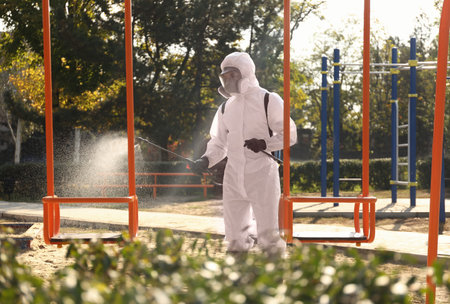Man in hazmat suit spraying disinfectant onto swing at children's playground.