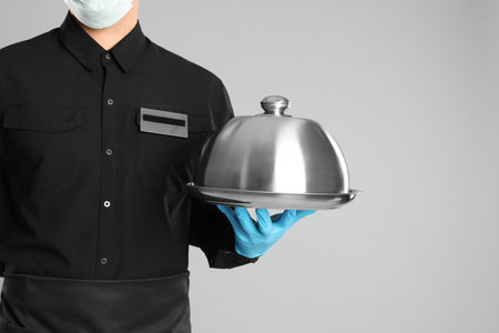 Waiter in medical face mask holding tray with lid on light gray background, closeupの写真素材