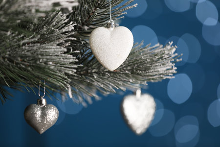 Beautiful holiday heart shaped baubles hanging on Christmas tree against blue background with blurred festive lightsの写真素材