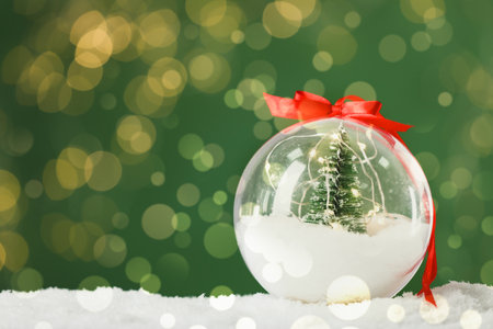 Beautiful transparent Christmas ornament with small fir tree and fairy lights on snow against green background, bokeh effect. Space for textの写真素材