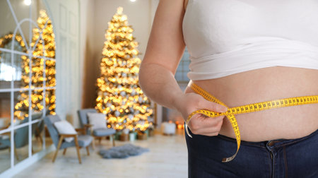 Overweight woman measuring her waist in room with Christmas tree after holidays, closeup