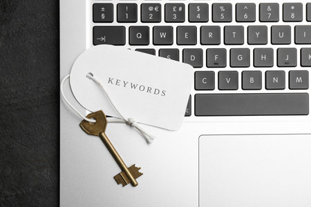 Laptop, key and tag with word KEYWORDS on black table, top view
