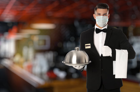 Waiter in medical face mask holding tray with lid in restaurantの写真素材