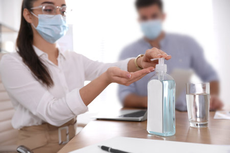 Office worker in protective mask using sanitizer at table, focus on hands. Personal hygiene during COVID-19 pandemicの写真素材