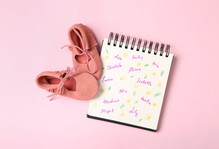 Notebook with different baby names and child's shoes on pink background, flat layの写真素材