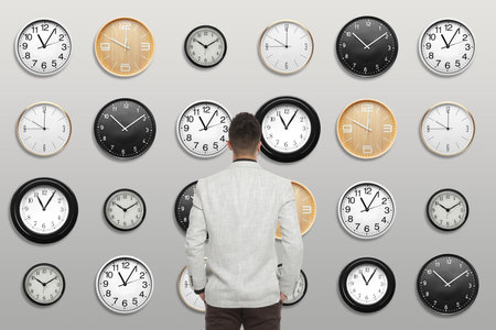 Time management concept. Businessman standing in front of different clocks on light background, back viewの写真素材